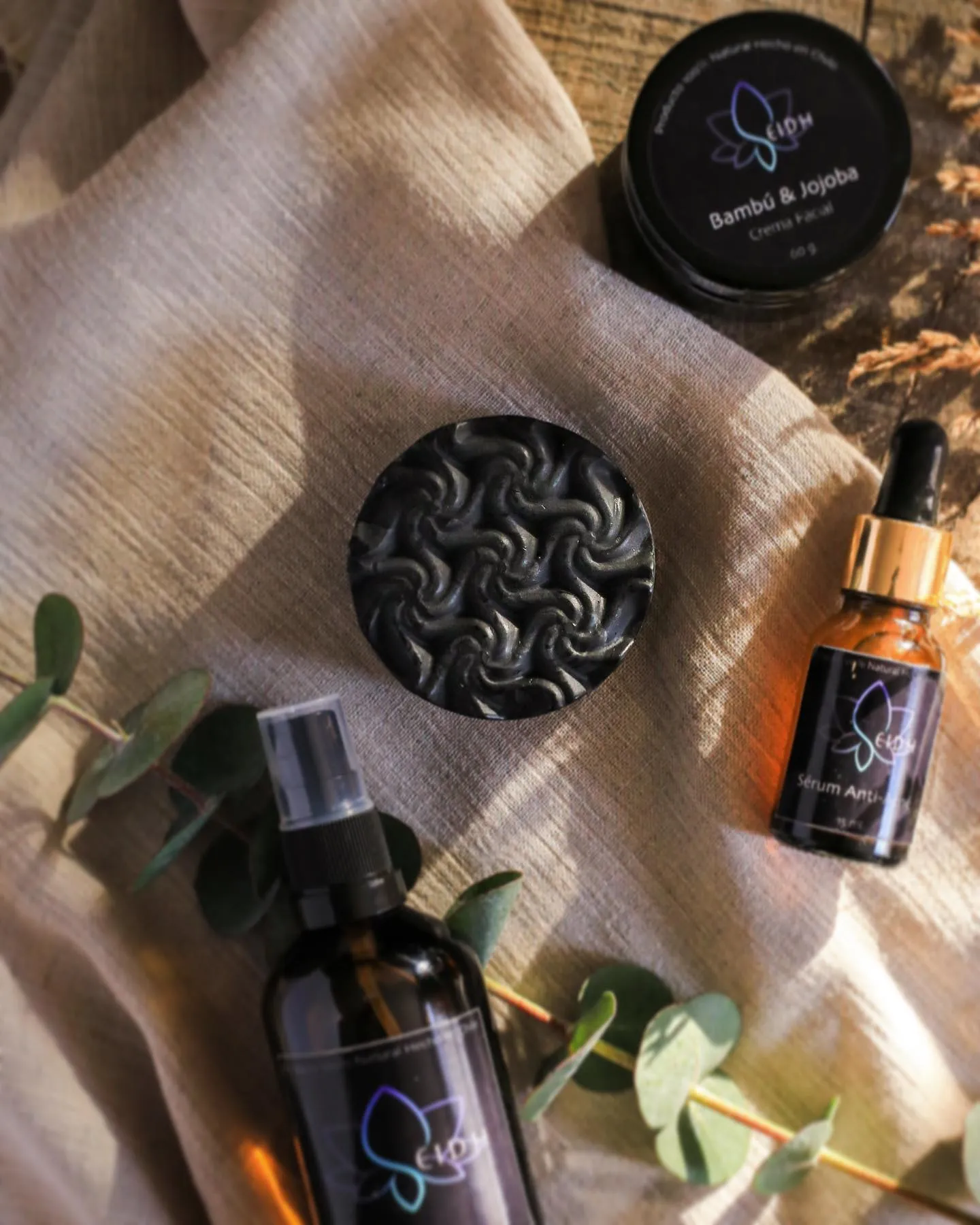 Composición de productos Seidh Cosmética (crema Bambú y Jojoba, jabón de carbón, sérum y bruma) sobre lino y madera con ramas de eucalipto